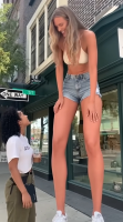 shrunken_woman giantesshug gentle city blonde long_hair clothed cleavage mini_giantess shorts bending_over laughing standing size_comparison bikini_top sidewalk ai_art